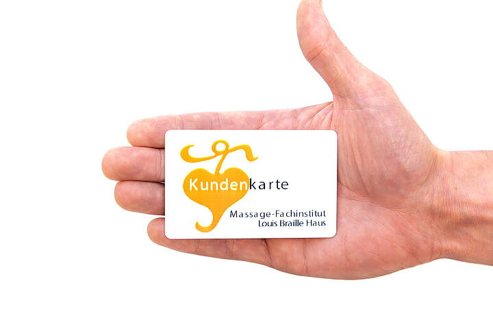 © BSVWNB/ Michael Schrenk Hand hält eine weiße Kundenkarte mit orangefarbenem Logo und der Aufschrift 'Massage-Fachinstitut Louis Braille Haus'