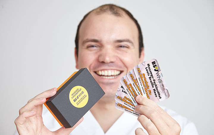 Masseur Sascha zeigt lächelnd Massage-Bons in die Kamera